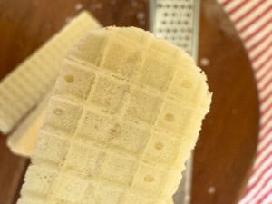 Close-up of a rectangular vanilla wafer cookie held above a brown surface with a metallic grater and red-striped fabric in the background.