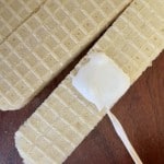 Three rectangular vanilla wafers on a wooden surface, with one wafer topped with a dollop of white cream and a small stick resting on it.