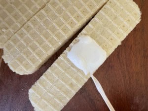 Three rectangular vanilla wafers on a wooden surface, with one wafer topped with a dollop of white cream and a small stick resting on it.