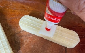 A hand squeezes red gel from a tube onto a square piece of white candy placed on top of a vanilla wafer cookie, all on a wooden surface.