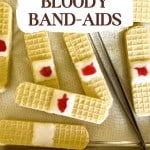 A baking tray with Bloody Band-Aids—wafer cookies decorated to look like band-aids, each topped with white frosting and a dab of red jelly resembling blood.