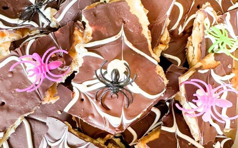 Chocolate-covered spider web saltine toffee bars topped with plastic toy spiders in black, pink, and green, arranged in a festive, Halloween-themed display.