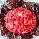 A molded, red gelatin salad shaped like a brain is placed on top of dark red lettuce leaves on a white towel.