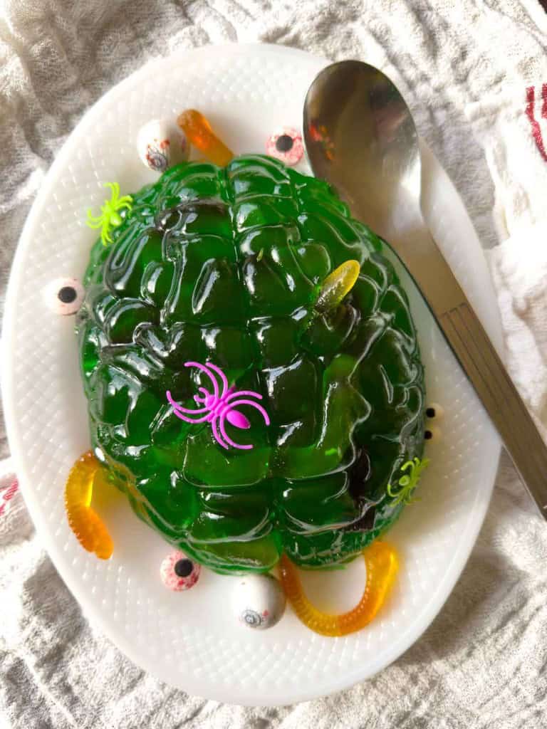 A green gelatin dessert shaped like a brain, decorated with gummy worms, candy eyeballs, and plastic spiders, on a white plate with a spoon.