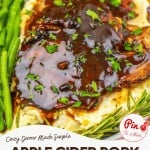 A plate with Apple Cider Pork Chops topped with savory sauce, served over creamy mashed potatoes, with green beans and fresh herbs on the side.