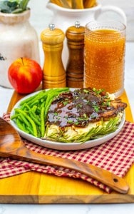 A plate of grilled chicken with sauce, mashed potatoes, and green beans sits on a red checkered cloth, next to an apple, salt and pepper shakers, a glass of iced tea, and savory Apple Cider Pork Chops.