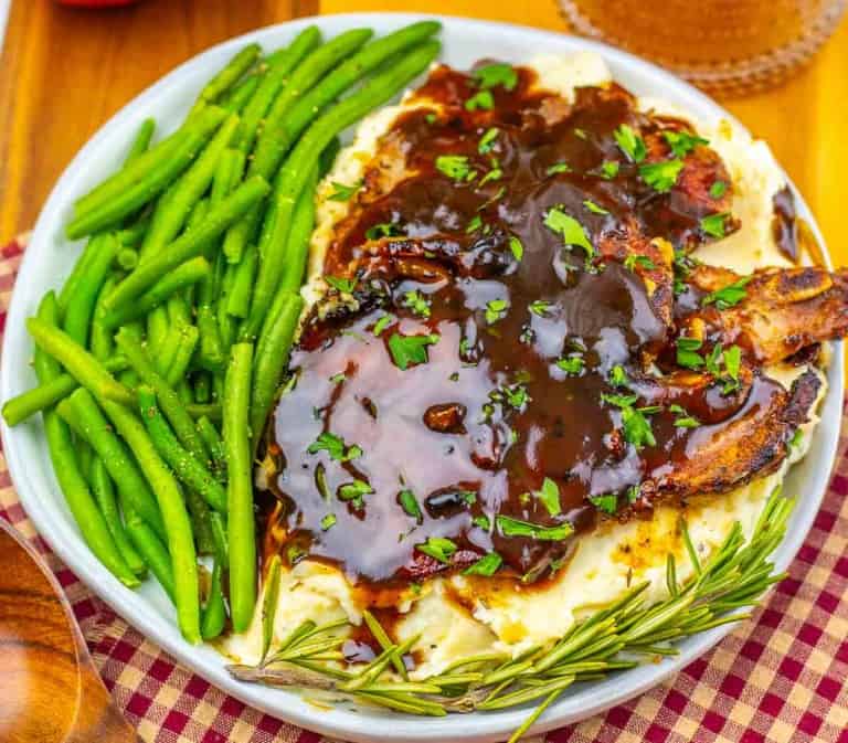 A plate of mashed potatoes topped with Apple Cider Pork Chops and brown gravy, served with green beans and garnished with fresh herbs and a sprig of rosemary.