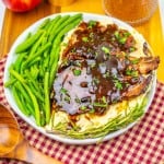 A plate of mashed potatoes topped with brown gravy and bacon, served alongside green beans and garnished with rosemary, pairs perfectly with savory Apple Cider Pork Chops on a wooden tray with a checkered cloth.