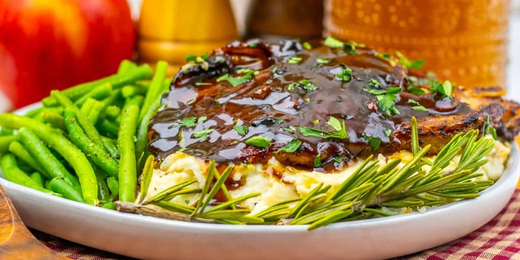 A plate of green beans, mashed potatoes, and Apple Cider Pork Chops topped with brown gravy, garnished with rosemary and chopped herbs.