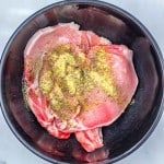 Raw pork chops in a black bowl, sprinkled with dried herbs and spices, ready to be transformed into delicious Apple Cider Pork Chops on a white marble surface.