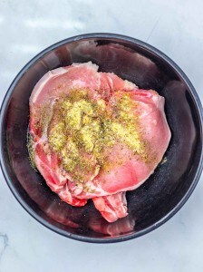 Raw pork chops in a black bowl, sprinkled with dried herbs and spices, ready to be transformed into delicious Apple Cider Pork Chops on a white marble surface.