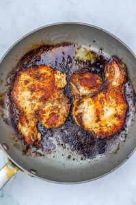 Two cooked, seasoned Apple Cider Pork Chops in a frying pan with browned bits and savory pan juices visible.