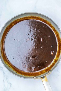 A stainless steel pan containing a thick, dark brown sauce with a glossy surface, perfect for drizzling over Apple Cider Pork Chops, shown from above on a light-colored countertop.