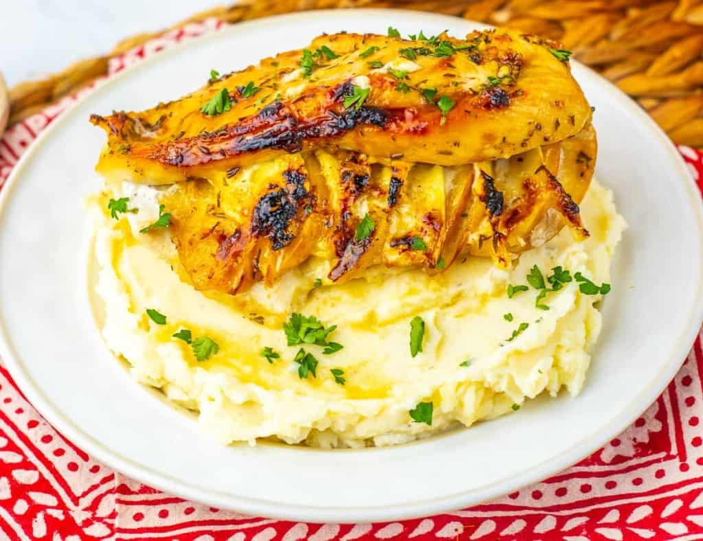 Grilled chicken breast served on a mound of mashed potatoes, garnished with chopped parsley, on a white plate.
