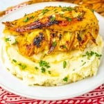 Grilled chicken breast served on a mound of mashed potatoes, garnished with chopped parsley, on a white plate.