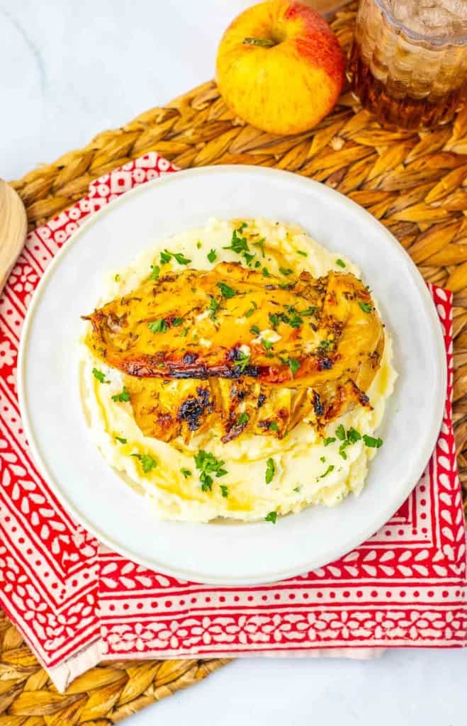 Grilled chicken breast served on mashed potatoes, garnished with chopped herbs, on a white plate with a red patterned napkin, an apple, and a glass of beverage nearby.