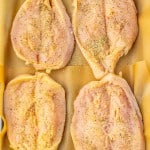 Four raw chicken breasts seasoned with spices are arranged on a baking sheet lined with parchment paper.