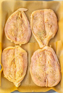 Four raw chicken breasts seasoned with spices are arranged on a baking sheet lined with parchment paper.