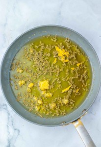 A frying pan on a marble surface contains oil, what appears to be minced garlic, small chunks of yellow paste, and scattered dried herbs or seeds.