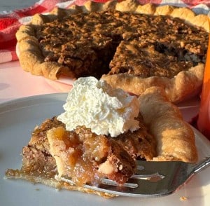 A slice of chopped pecan pie with a crumbly filling and whipped cream on top is served on a plate in front of the whole pie with a missing slice.