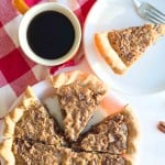 A sliced chopped pecan Pie on a plate with one piece served on a separate plate and a cup of black coffee on a red and white checkered tablecloth.