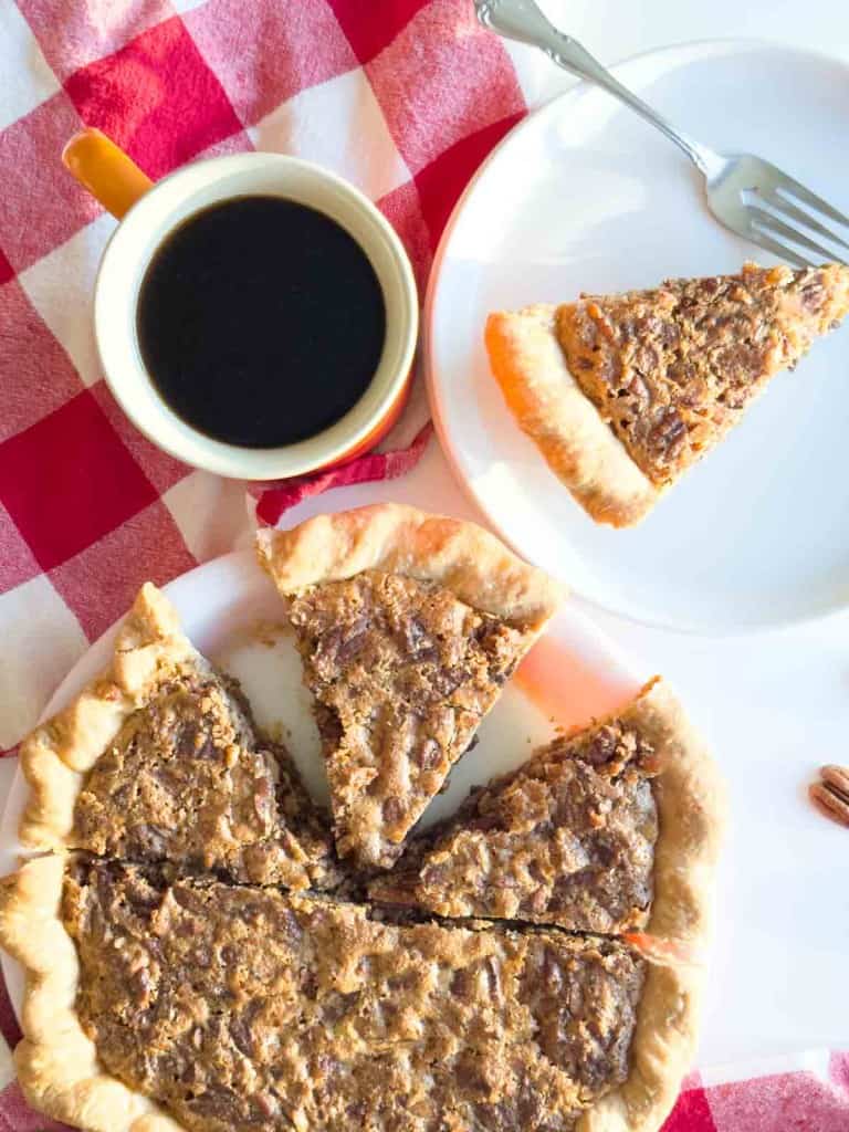 A sliced chopped pecan Pie on a plate with one piece served on a separate plate and a cup of black coffee on a red and white checkered tablecloth.