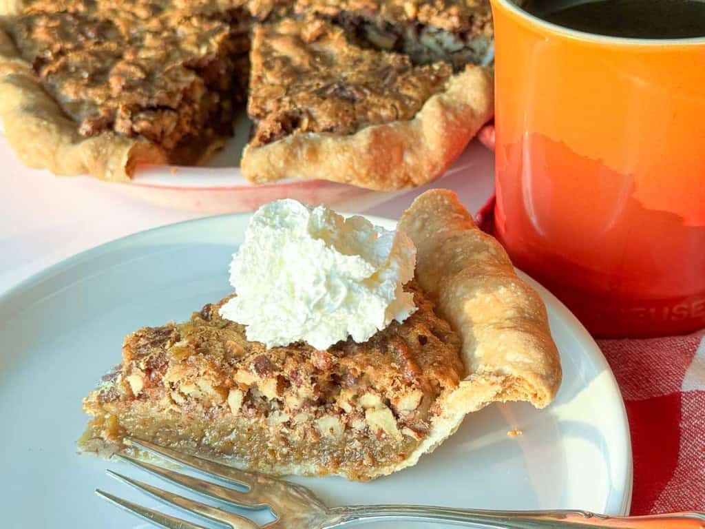 A slice of chopped pecan pie topped with whipped cream sits on a white plate with a fork, next to an orange mug and a full pecan pie in the background.