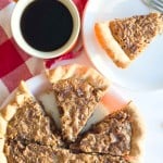 Chopped pecan pie slices rest on a white plate, paired with a cup of black coffee and a red and white checkered napkin nearby.