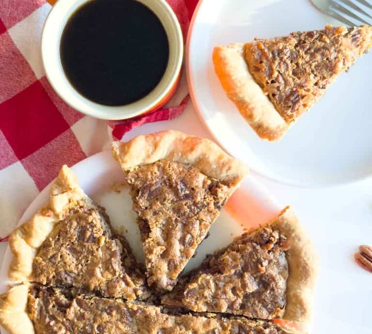 Chopped pecan pie slices rest on a white plate, paired with a cup of black coffee and a red and white checkered napkin nearby.