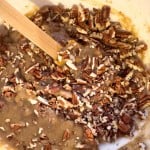 Chopped pecans being mixed into a thick, brown batter with a wooden spatula in a white bowl.