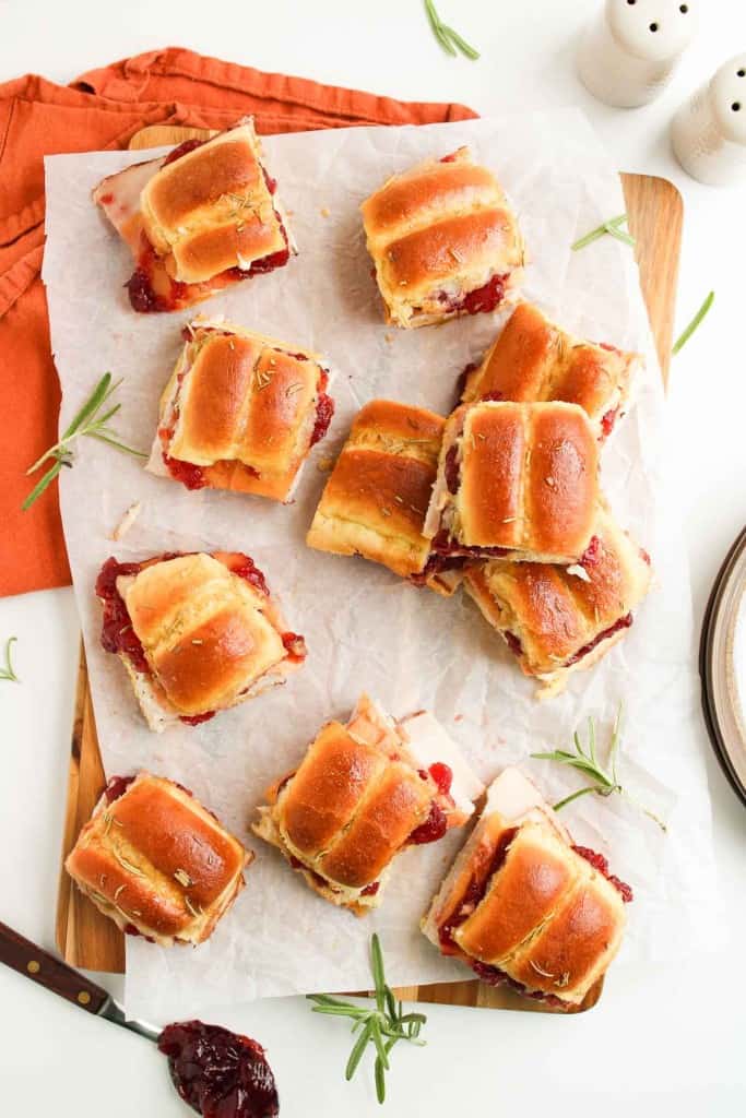 Twelve mini sandwiches with ham, cheese, and cranberry sauce on a parchment-lined board, garnished with rosemary.