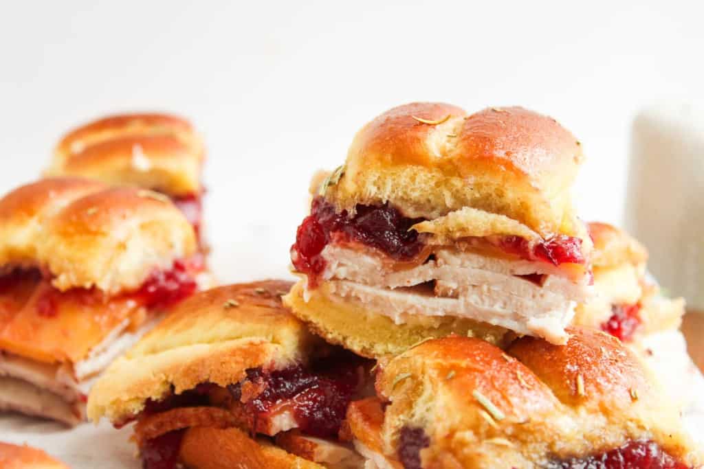 A close-up of small turkey sandwiches with cranberry sauce on soft rolls, stacked on a white surface.