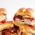 A close-up of small turkey sandwiches with cranberry sauce on soft rolls, stacked on a white surface.