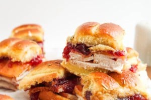 A close-up of small turkey sandwiches with cranberry sauce on soft rolls, stacked on a white surface.