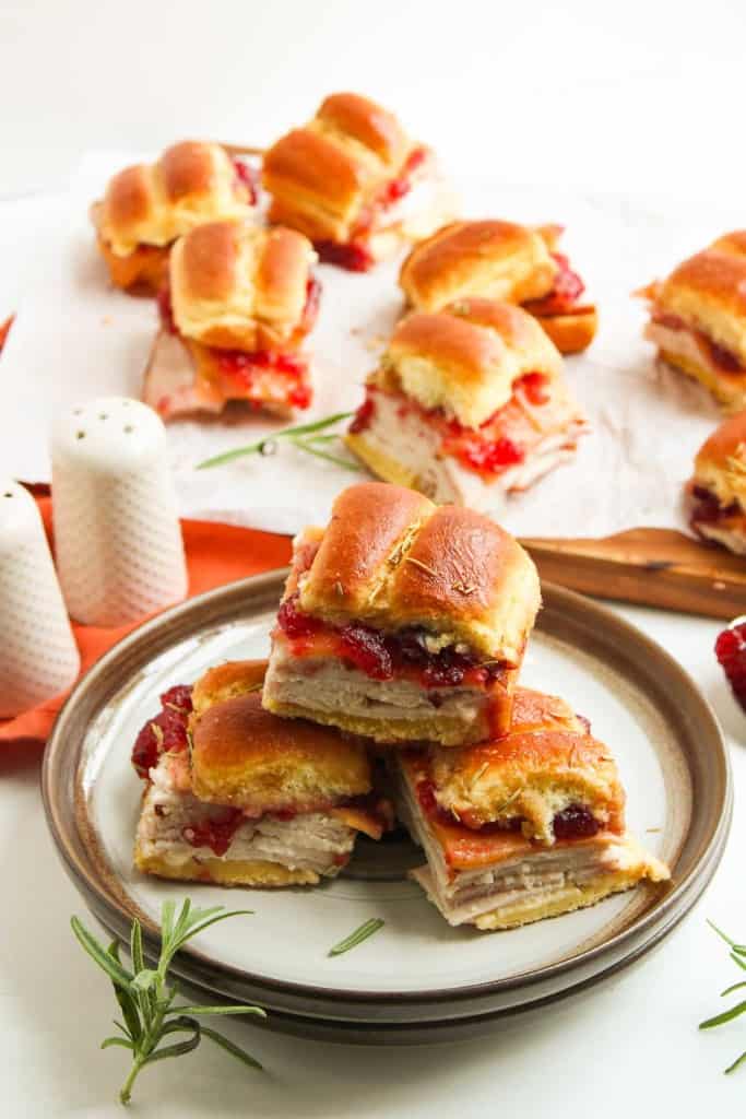 Three slider sandwiches with turkey and cranberry sauce are stacked on a plate, with more sandwiches in the background on parchment paper. Salt and pepper shakers and rosemary sprigs are nearby.