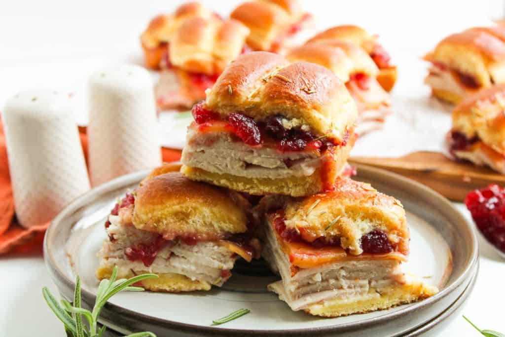 Three small sandwiches with turkey, cranberry sauce, and soft rolls are stacked on a plate. More sandwiches are visible in the background beside salt and pepper shakers.
