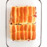 A glass baking dish containing twelve golden brown slider sandwiches, arranged in a 3x4 grid, on a white background.