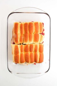 A glass baking dish containing twelve golden brown slider sandwiches, arranged in a 3x4 grid, on a white background.