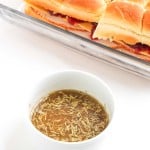 A white bowl of seasoned au jus sits in front of a glass dish filled with stacked deli meat sliders on soft rolls.