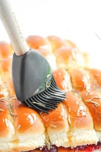 A silicone pastry brush is spreading glaze over a batch of shiny, golden-brown dinner rolls sprinkled with herbs.