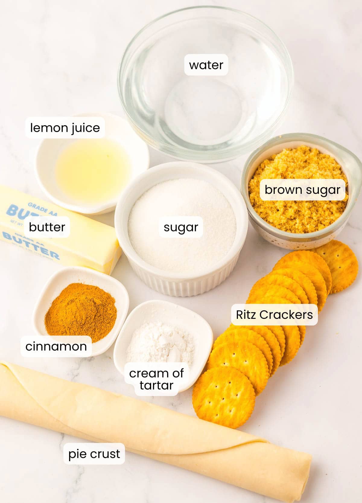 Ingredients for a ritz cracker pie are arranged on a countertop, including pie crust, Ritz crackers, butter, sugar, brown sugar, water, lemon juice, cinnamon, and cream of tartar.