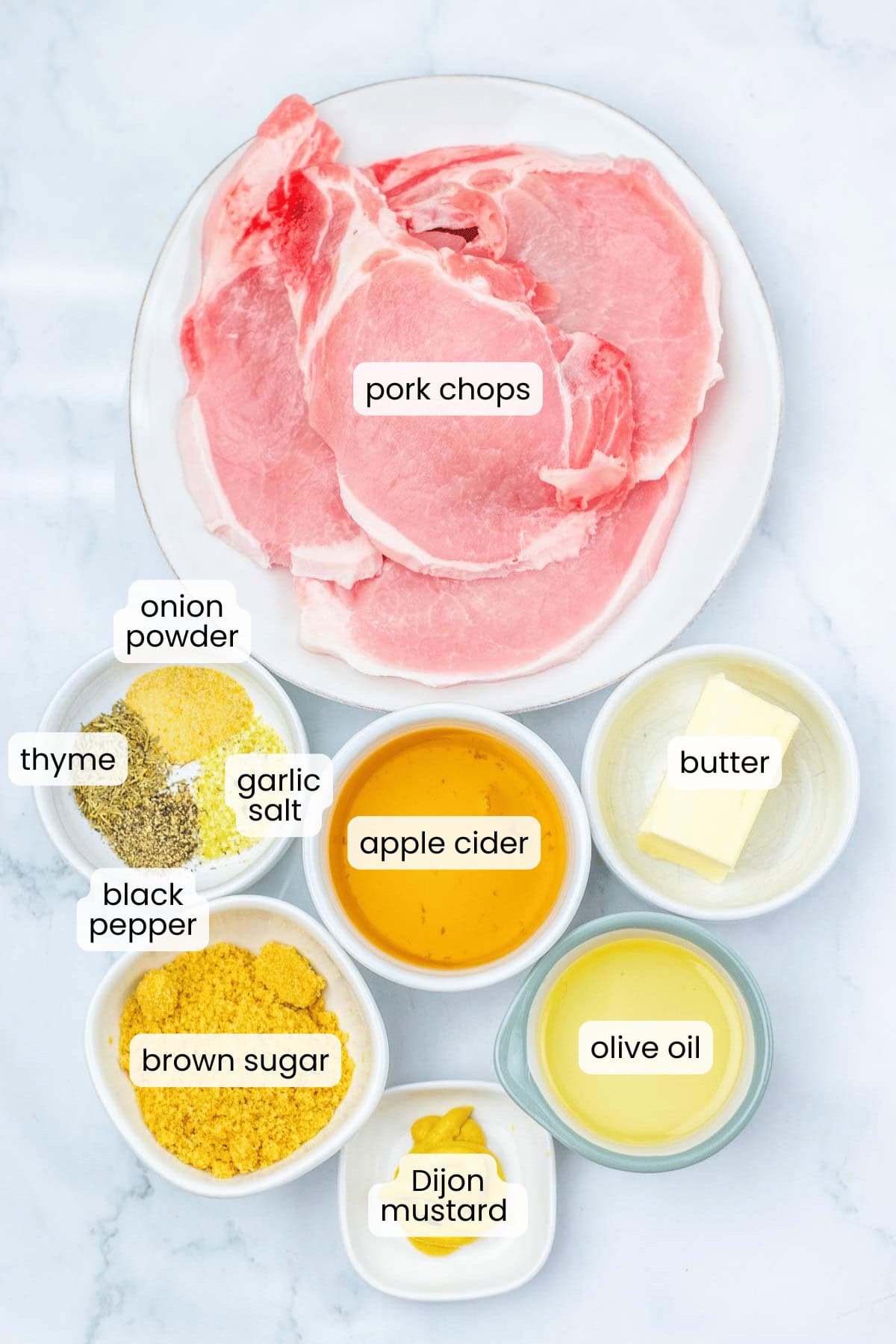 A plate of raw Apple Cider Pork Chops is surrounded by small bowls of onion powder, garlic salt, thyme, black pepper, brown sugar, apple cider, Dijon mustard, olive oil, and butter.
