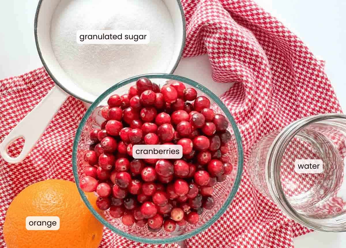 A bowl of cranberries, a measuring cup of granulated sugar, an orange, and a jar of water—everything you need to make homemade orange cranberry sauce—are arranged on a red and white checkered cloth.