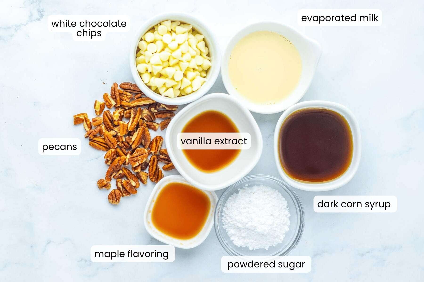 Six labeled ingredients for pecan pie fudge on a white surface: white chocolate chips, evaporated milk, dark corn syrup, powdered sugar, maple flavoring, pecans, and vanilla extract.