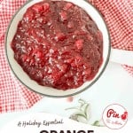 A bowl of chunky orange cranberry sauce sits on a white surface with a red and white checkered cloth beside it. Text on the image reads "Orange Cranberry Sauce," highlighting this festive, homemade orange cranberry sauce.