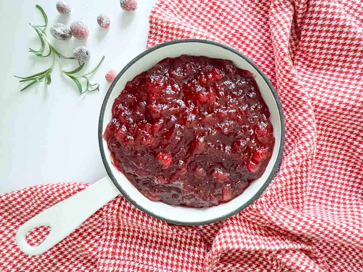 Skillet of orange cranberry sauce on a red houndstooth cloth with sugared cranberries and rosemary beside it.