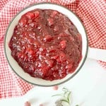 A white bowl filled with orange cranberry sauce sits on a white surface, surrounded by a red and white checkered cloth, sugared cranberries, and fresh rosemary.