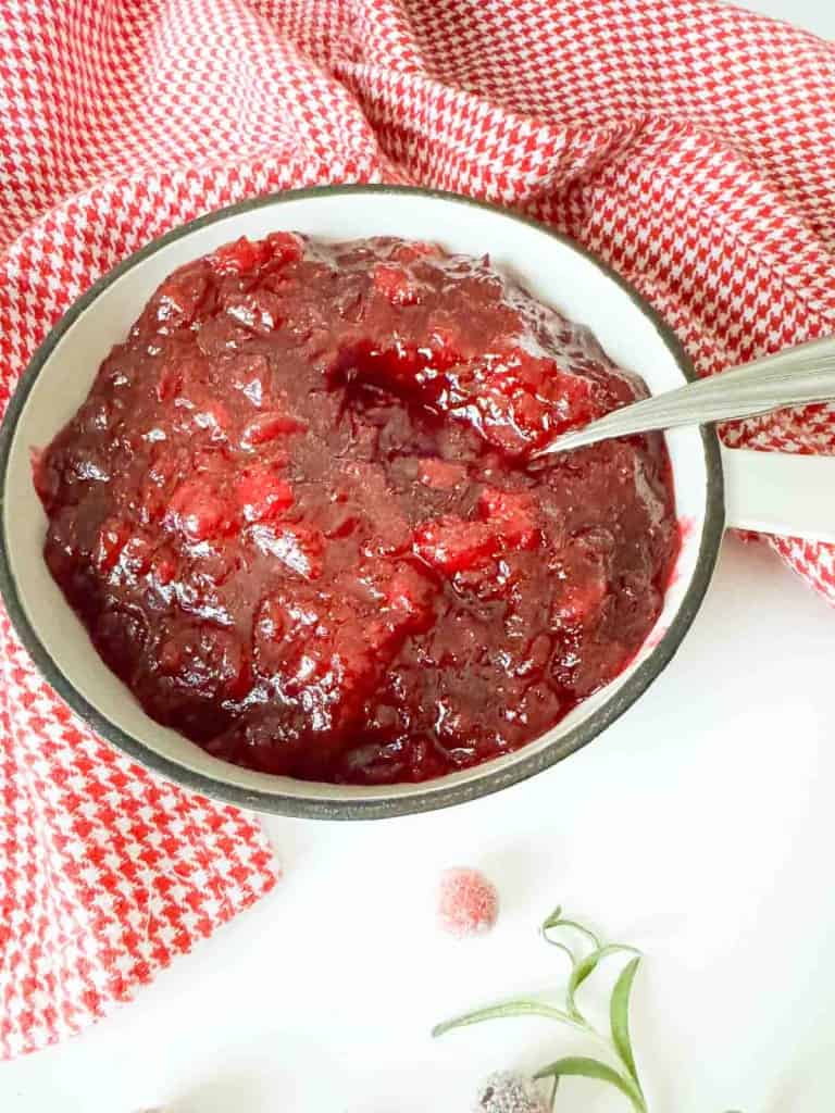 A white bowl filled with orange cranberry sauce sits on a white surface next to a red and white checkered cloth, with a spoon in the bowl.