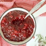 A white bowl filled with chunky orange cranberry sauce sits on a red and white checkered cloth, with a spoon in the bowl and sprigs of rosemary nearby.