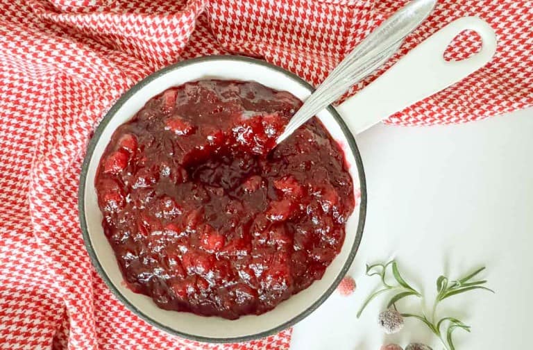A white bowl filled with chunky orange cranberry sauce sits on a red and white checkered cloth, with a spoon in the bowl and sprigs of rosemary nearby.
